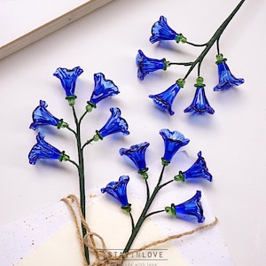 May include: Two stems of blue glass flowers with green detailing. The bell-shaped flowers are arranged on green stems. The stems are placed on a white surface with a card and twine. The text "STAY IN LOVE" is visible.