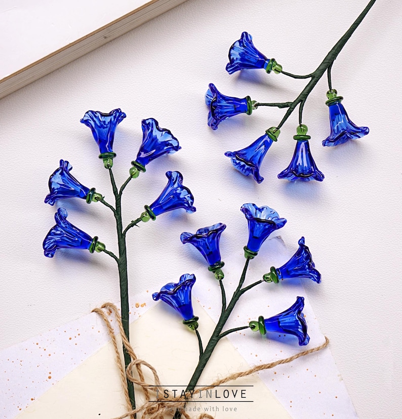 May include: Two stems of blue glass flowers with green detailing. The bell-shaped flowers are arranged on green stems. The stems are placed on a white surface with a card and twine. The text "STAY IN LOVE" is visible.