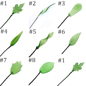 May include: Nine green glass leaf stems, each with a different leaf design. The leaves vary in shape, from simple ovals to more complex, textured forms. Each leaf is attached to a dark green or black wire stem.