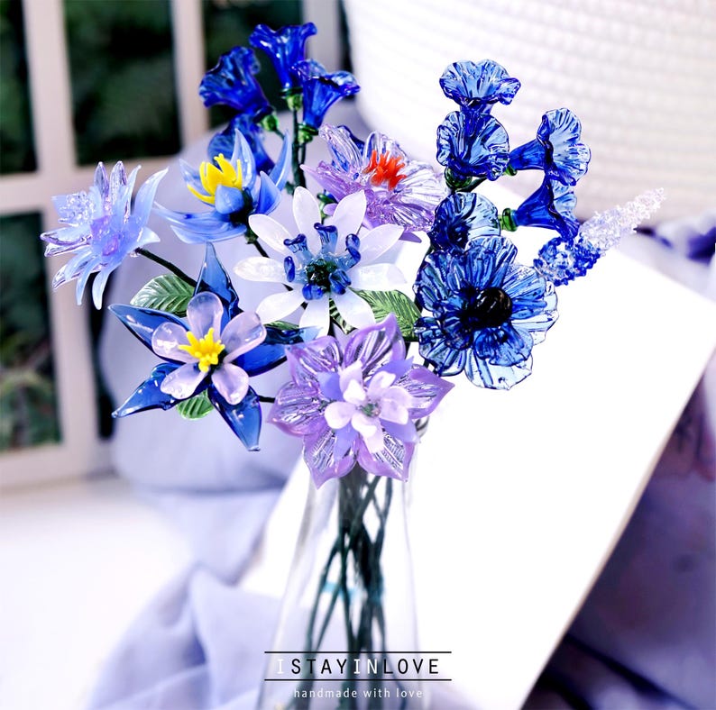 May include: A bouquet of handcrafted glass flowers in a clear vase. The flowers are various shades of blue, purple, white, and yellow. The vase sits on a white surface. The text "I STAY IN LOVE handmade with love" is at the bottom.
