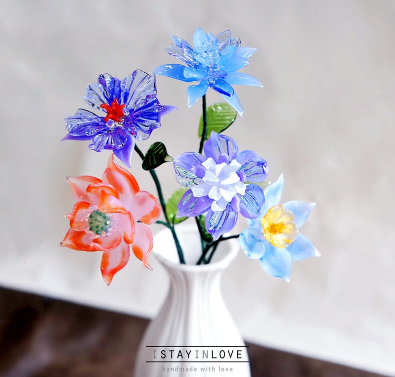 May include: A bouquet of colorful glass flowers in a white vase. The flowers are various shades of blue, purple, and orange, with green stems and leaves. The vase has the text "I STAY IN LOVE" at the bottom.