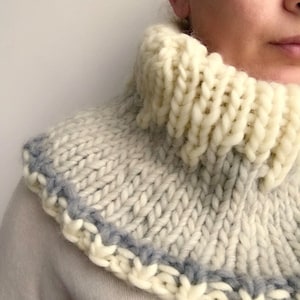 May include: A white knitted cowl neck sweater with a blue trim. The cowl neck is thick and fluffy.