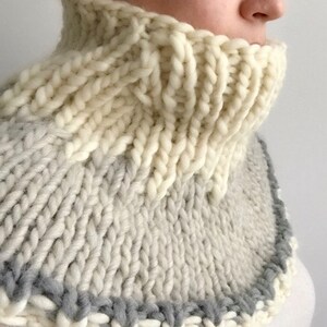 May include: A white and gray knitted cowl neck warmer with a gray trim. The cowl is made of thick yarn and has a chunky knit texture.