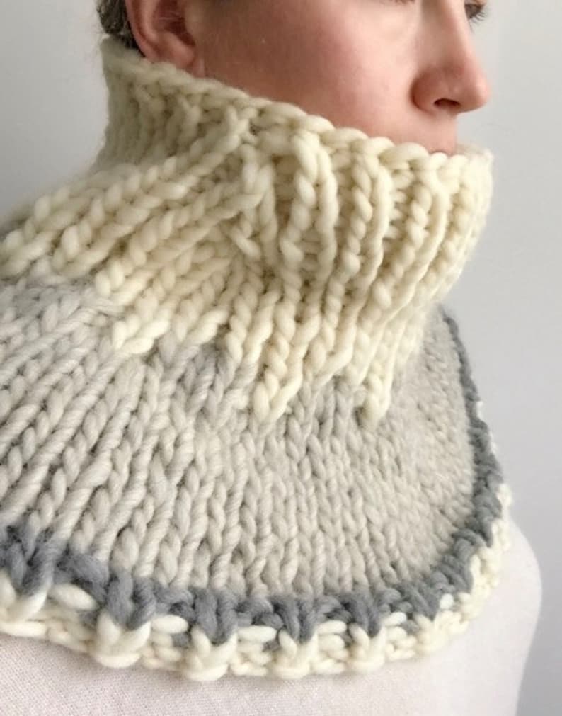May include: A white and gray knitted cowl neck warmer with a gray trim. The cowl is made of thick yarn and has a chunky knit texture.
