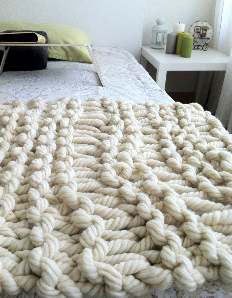 May include: A close-up of a chunky knit throw blanket in a cream color. The blanket is made of thick yarn and has a textured, cable knit pattern.