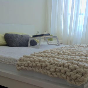 May include: A white bed with a white knitted blanket, two pillows, and a small white table with a book on it. The bed is in a bedroom with a window and white curtains.
