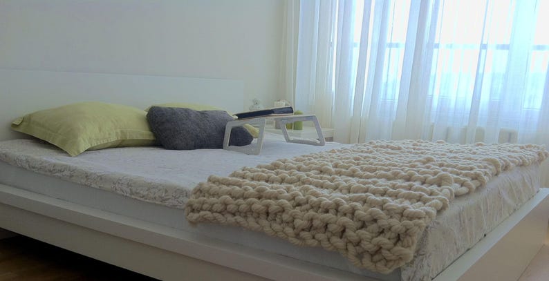 May include: A white bed with a white knitted blanket, two pillows, and a small white table with a book on it. The bed is in a bedroom with a window and white curtains.
