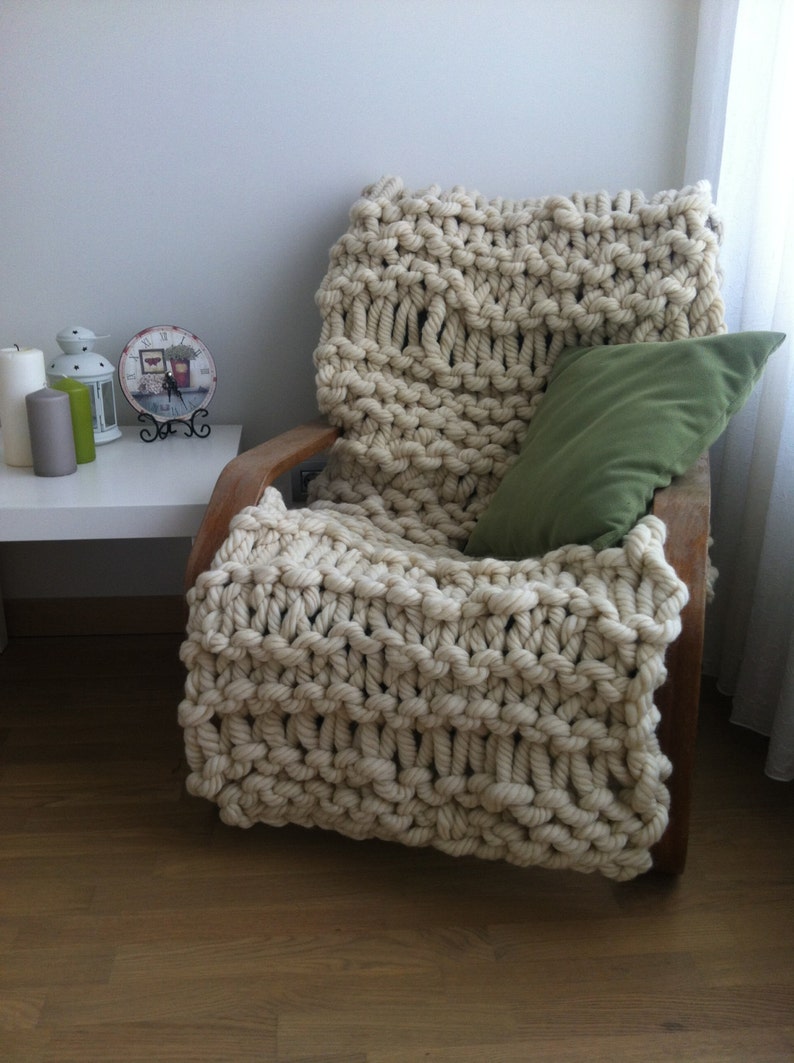 May include: A large, chunky knit throw blanket in a cream color is draped over a wooden armchair. A green pillow is placed on the chair. The blanket has a textured, cable knit pattern.