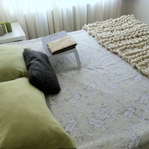 May include: A white bed with a floral patterned sheet and a white knitted throw blanket. There are two green pillows and one gray pillow on the bed. A wooden tray with a book is on the bed.