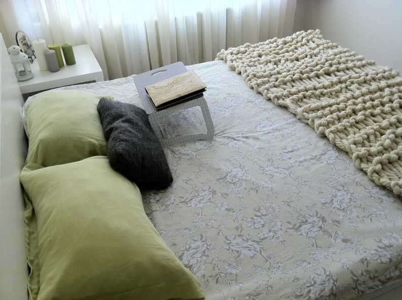 May include: A white bed with a floral patterned sheet and a white knitted throw blanket. There are two green pillows and one gray pillow on the bed. A wooden tray with a book is on the bed.