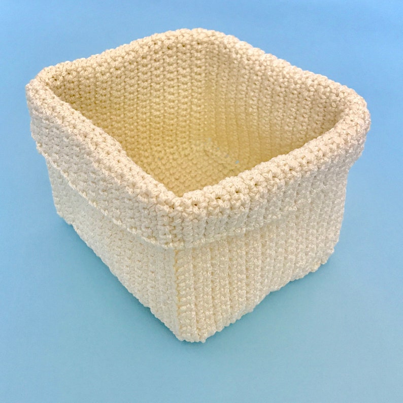May include: A white crocheted basket with a square shape. The basket is made with a thick yarn and has a woven texture.