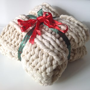 Op de afbeelding: Een grote, cr&egrave;mekleurige gebreide deken met een rood en groen lint dat bovenop in een strik is gebonden. Het lint heeft de tekst "Merry Christmas" erop gedrukt.