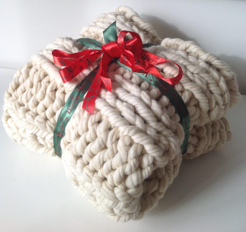 Op de afbeelding: Een grote, cr&egrave;mekleurige gebreide deken met een rood en groen lint dat bovenop in een strik is gebonden. Het lint heeft de tekst "Merry Christmas" erop gedrukt.