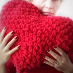 May include: A large, heart-shaped, red knitted pillow. The pillow is made of a chunky, textured yarn. The image shows the pillow being held, with the hands of the person holding it visible. The nails are painted red.
