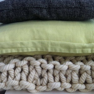 May include: A stack of three pillows, the top one is dark gray, the middle one is light green, and the bottom one is a cream-colored knitted throw pillow.