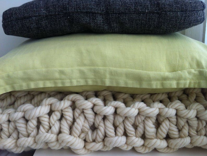 May include: A stack of three pillows, the top one is dark gray, the middle one is light green, and the bottom one is a cream-colored knitted throw pillow.