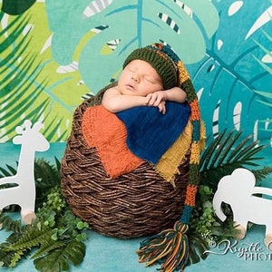 May include: A newborn baby wearing a green, yellow, and orange crocheted hat with a long tassel sleeps in a woven basket. The baby is surrounded by green foliage and white wooden animal toys, including a giraffe and an elephant.