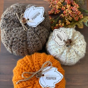 Puede incluir: Tres calabazas de crochet en marr&oacute;n, blanco y naranja con etiquetas en forma de hoja que dicen "Handmade Pumpkin".