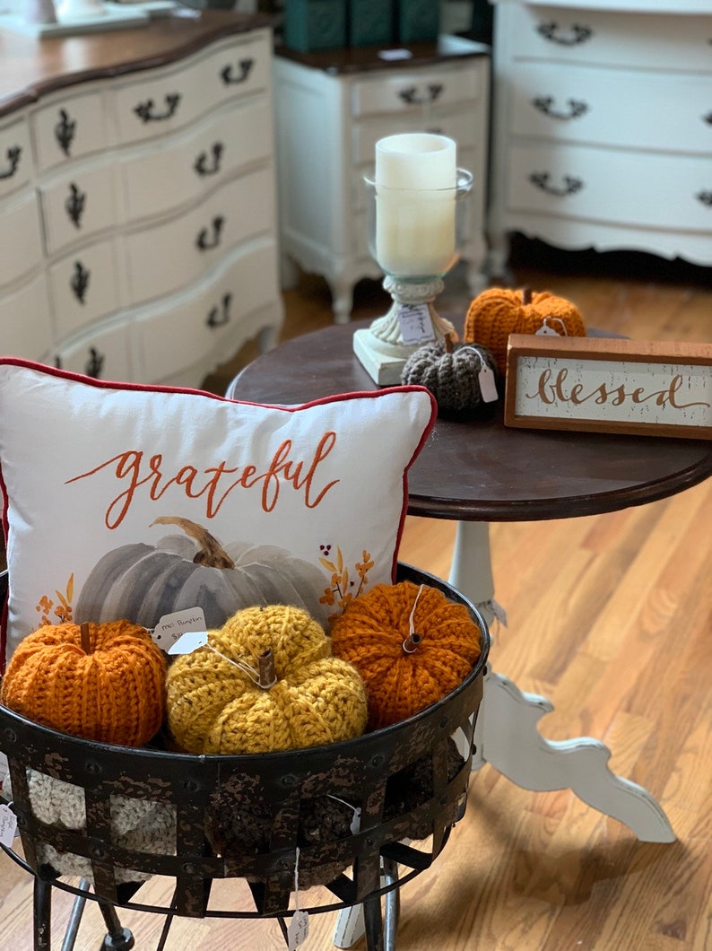 Puede incluir: Un soporte de metal negro con calabazas de crochet en naranja y amarillo. El soporte est&aacute; decorado con una almohada que dice "grateful" y un letrero que dice "blessed".