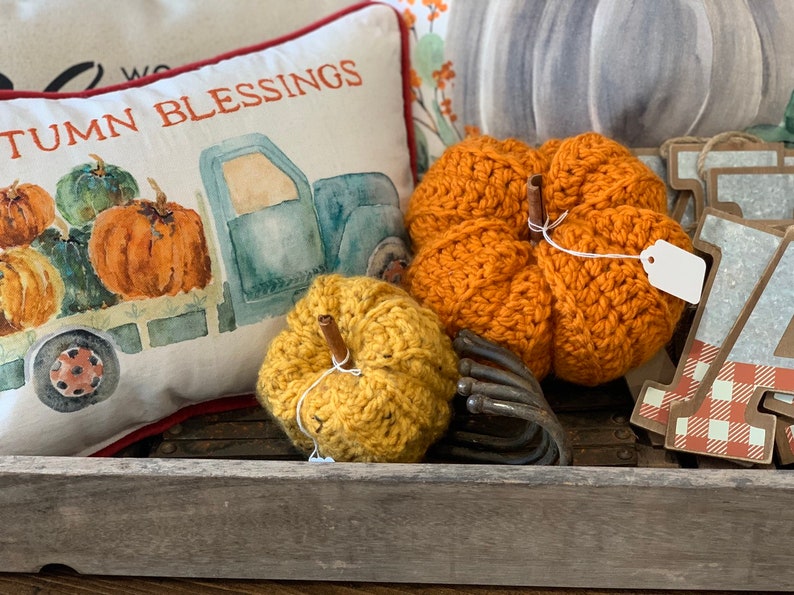 Puede incluir: Dos calabazas de crochet, una naranja y otra amarilla, con tallos de madera, sentadas en una bandeja de madera con una almohada que muestra un cami&oacute;n que lleva calabazas y el texto "Autumn Blessings".
