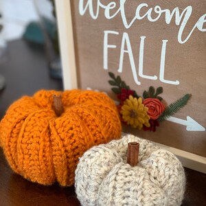 Puede incluir: Dos calabazas de crochet, una naranja y otra blanca, con tallos de canela. Las calabazas est&aacute;n sobre una superficie de madera delante de un cartel que dice "Bienvenido Oto&ntilde;o" con un dise&ntilde;o floral.