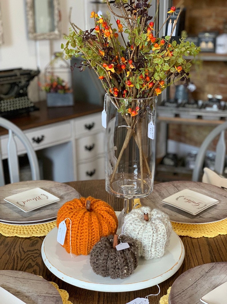 Puede incluir: Tres calabazas de crochet en naranja, blanco y marr&oacute;n sobre un soporte para pasteles blanco. Las calabazas est&aacute;n sobre una mesa de madera con manteles individuales de crochet amarillos. Un jarr&oacute;n con follaje de oto&ntilde;o est&aacute; en el fondo.