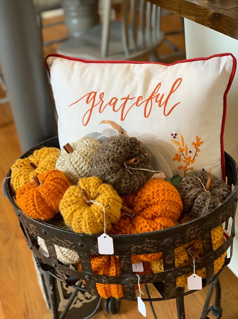 Puede incluir: Una cesta de metal llena de calabazas de crochet en varios colores, incluyendo naranja, amarillo, marr&oacute;n y crema. La cesta est&aacute; sentada en una mesa con una almohada blanca que dice "agradecido" en letras naranjas.