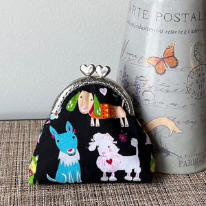 May include: A black coin purse with a silver clasp, featuring a whimsical dog print in various colors. The purse is next to a vase with flowers and the text "ARTE POSTALE".