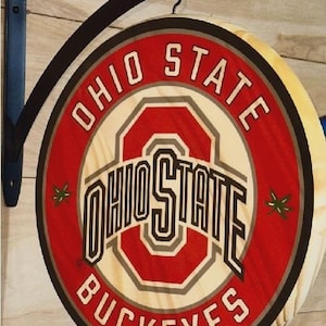 May include: Round wooden sign featuring the Ohio State Buckeyes logo in red, white, and gray. The sign is mounted on a black metal bracket. The sign reads "OHIO STATE BUCKEYES" in a circular design.
