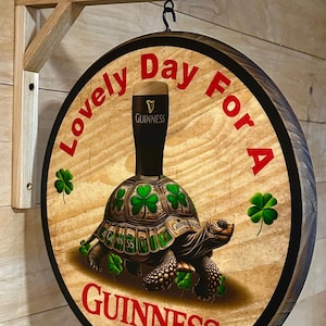 May include: A round wooden sign with a black border and a brown woodgrain background. The sign features a green turtle with a glass of Guinness beer on its back. The text "Lovely Day For A Guinness" is written in red letters around the turtle.