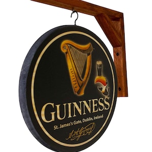 May include: A round wooden sign with a black background and a gold Guinness harp logo. The sign also features the text "GUINNESS", "St. James's Gate, Dublin, Ireland", and a signature. The sign is hanging from a wooden bracket.