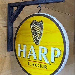 May include: A round wooden sign with a black border and a yellow background. The sign features a harp graphic and the text "HARP LAGER". The sign is hanging from a black metal bracket.