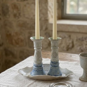 Può includere: Due candele affusolate, alte e sottili, di colore bianco sporco, sono posizionate verticalmente in una coppia di candelabri in ceramica abbinati. I supporti presentano uno smalto crema maculato che sfuma in una base azzurra decorata.