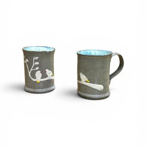 May include: Two cylindrical ceramic mugs with a dark gray speckled exterior and a light blue interior glaze. Each mug features a hand-painted white branch design with small white birds perched on the side, accented with yellow and red details.