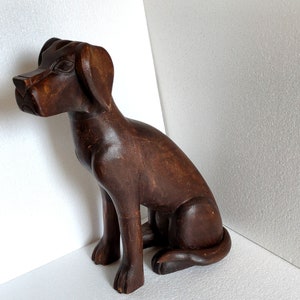 May include: A dark brown wooden sculpture of a dog sitting with its head turned to the side. The dog has a detailed face and a long tail.