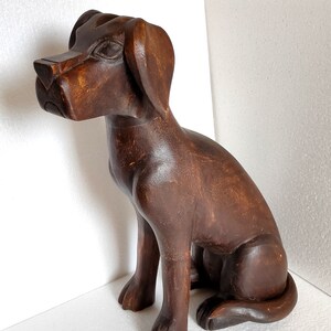 May include: A dark brown wooden sculpture of a dog sitting with its head tilted to the side. The dog has a detailed face and a long tail.