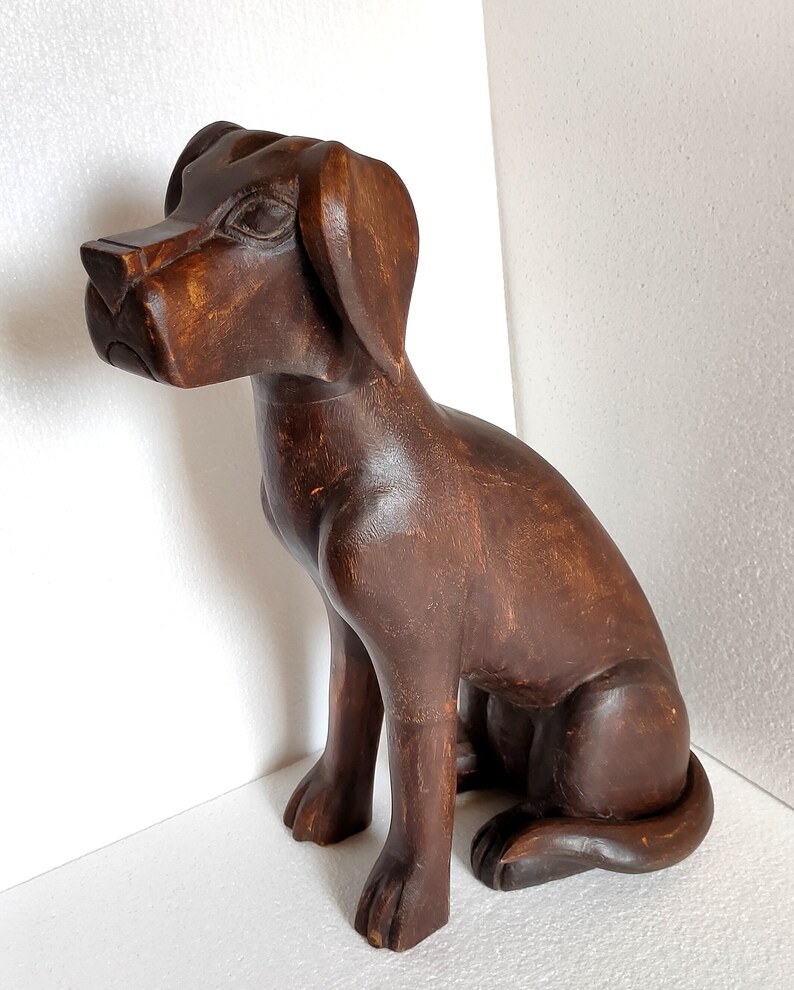 May include: A dark brown wooden sculpture of a dog sitting with its head tilted to the side. The dog has a detailed face and a long tail.