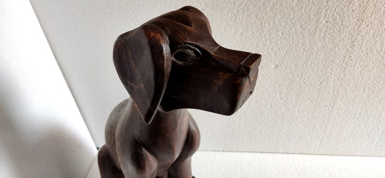 May include: A dark brown wooden sculpture of a dog sitting with its head tilted to the side. The dog has a long snout and large ears.