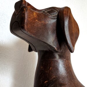 May include: A dark brown wooden sculpture of a dog with its head tilted upwards. The sculpture is carved in a stylised, abstract style.