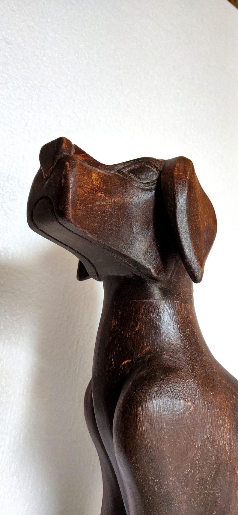 May include: A dark brown wooden sculpture of a dog with its head tilted upwards. The sculpture is carved in a stylised, abstract style.