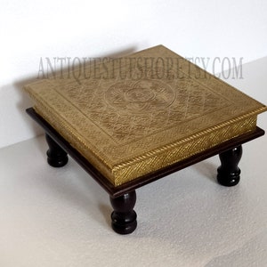 May include: A square brass table with an intricate floral design on the top and four dark wood legs. The table is approximately 18 inches wide and 18 inches long.