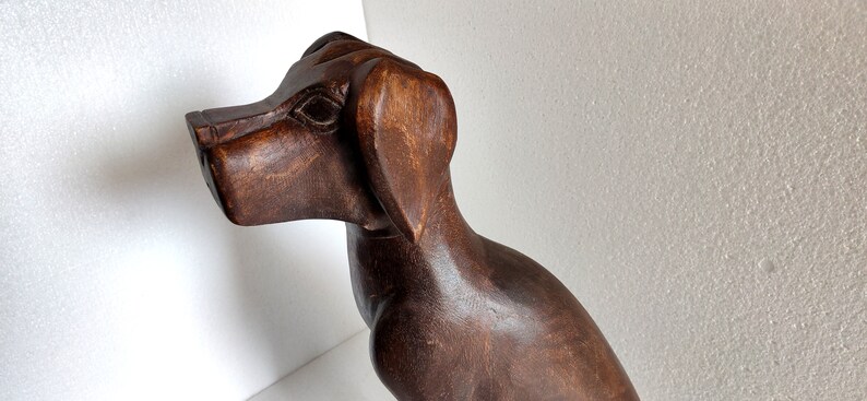 May include: A dark brown wooden sculpture of a dog. The dog is sitting with its head turned to the side and its ears perked up.