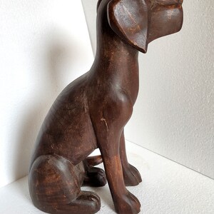May include: A dark brown wooden sculpture of a dog sitting with its head turned to the right. The dog has a long snout and large ears.