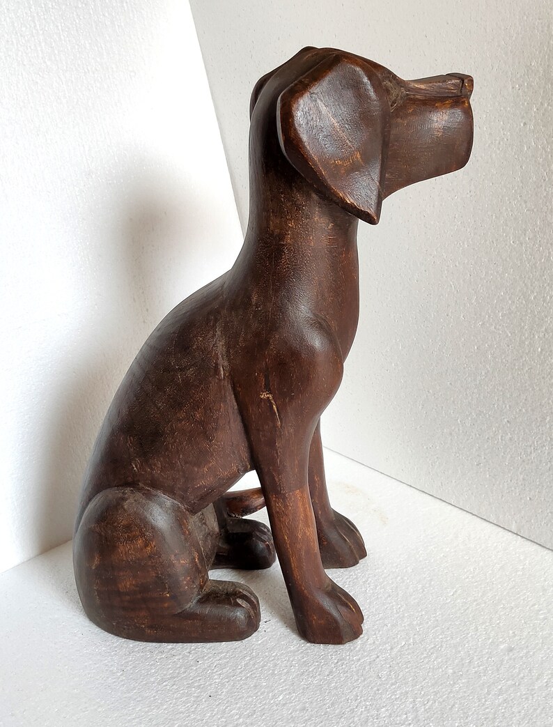 May include: A dark brown wooden sculpture of a dog sitting with its head turned to the right. The dog has a long snout and large ears.