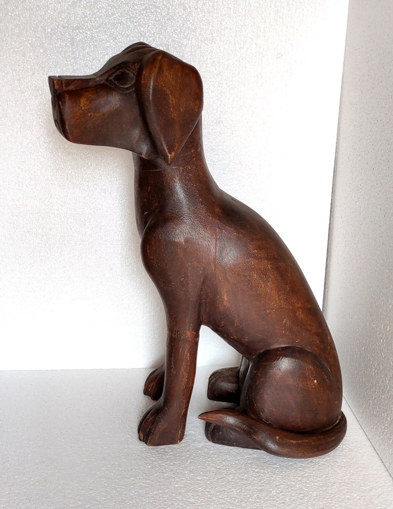 May include: A dark brown wooden sculpture of a dog sitting with its head turned to the side. The dog has a long snout and floppy ears.