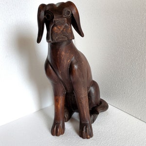 May include: A dark brown wooden sculpture of a dog sitting with its head tilted slightly to the side. The dog has a long snout and large ears.