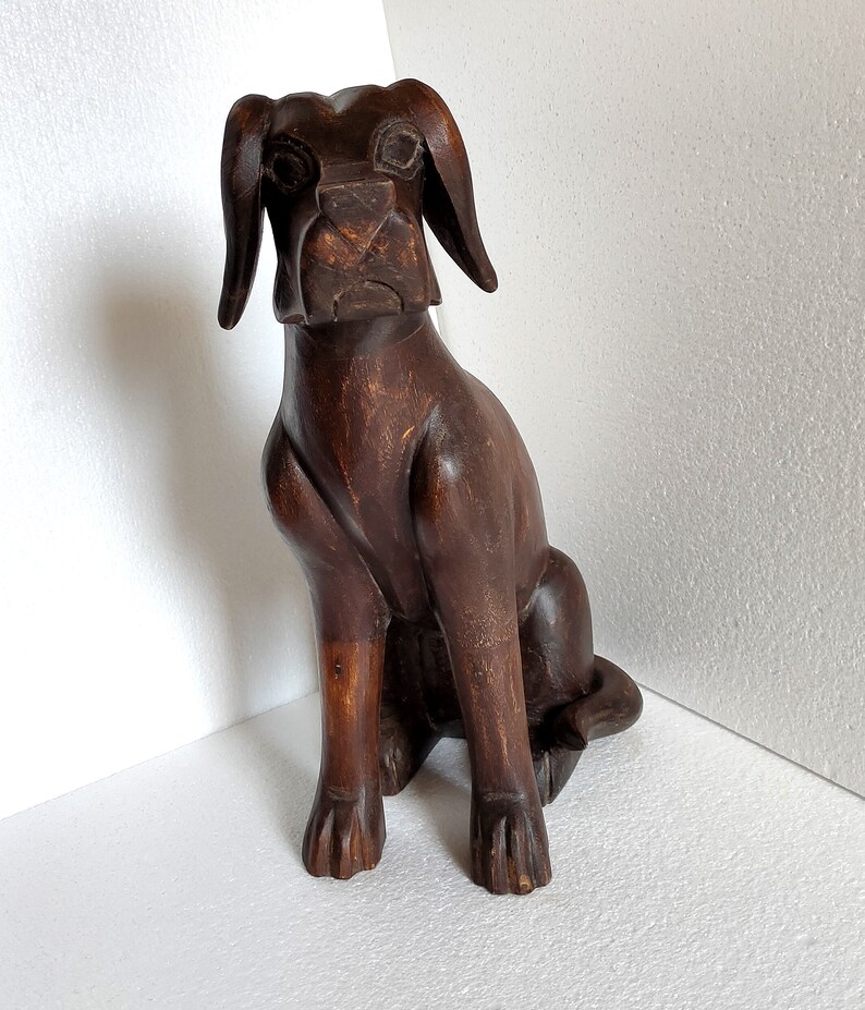 May include: A dark brown wooden sculpture of a dog sitting with its head tilted slightly to the side. The dog has a long snout and large ears.