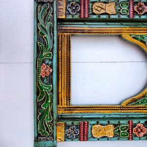 May include: A decorative wooden frame with intricate carvings and colorful paint. The frame is painted in shades of green, yellow, red, and blue. The frame has a floral design and a geometric pattern.