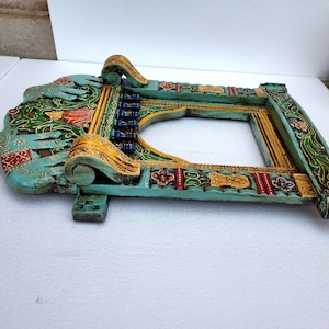 May include: A teal blue wooden frame with intricate carvings and colorful paint details. The frame features a carved archway design with floral and elephant motifs.