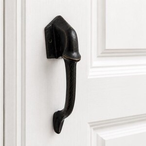 Peut inclure: Une poignée de porte en bronze foncé montée sur une porte blanche. La poignée a un design incurvé et texturé et une plaque de montage rectangulaire. La porte a un design à panneaux en relief.